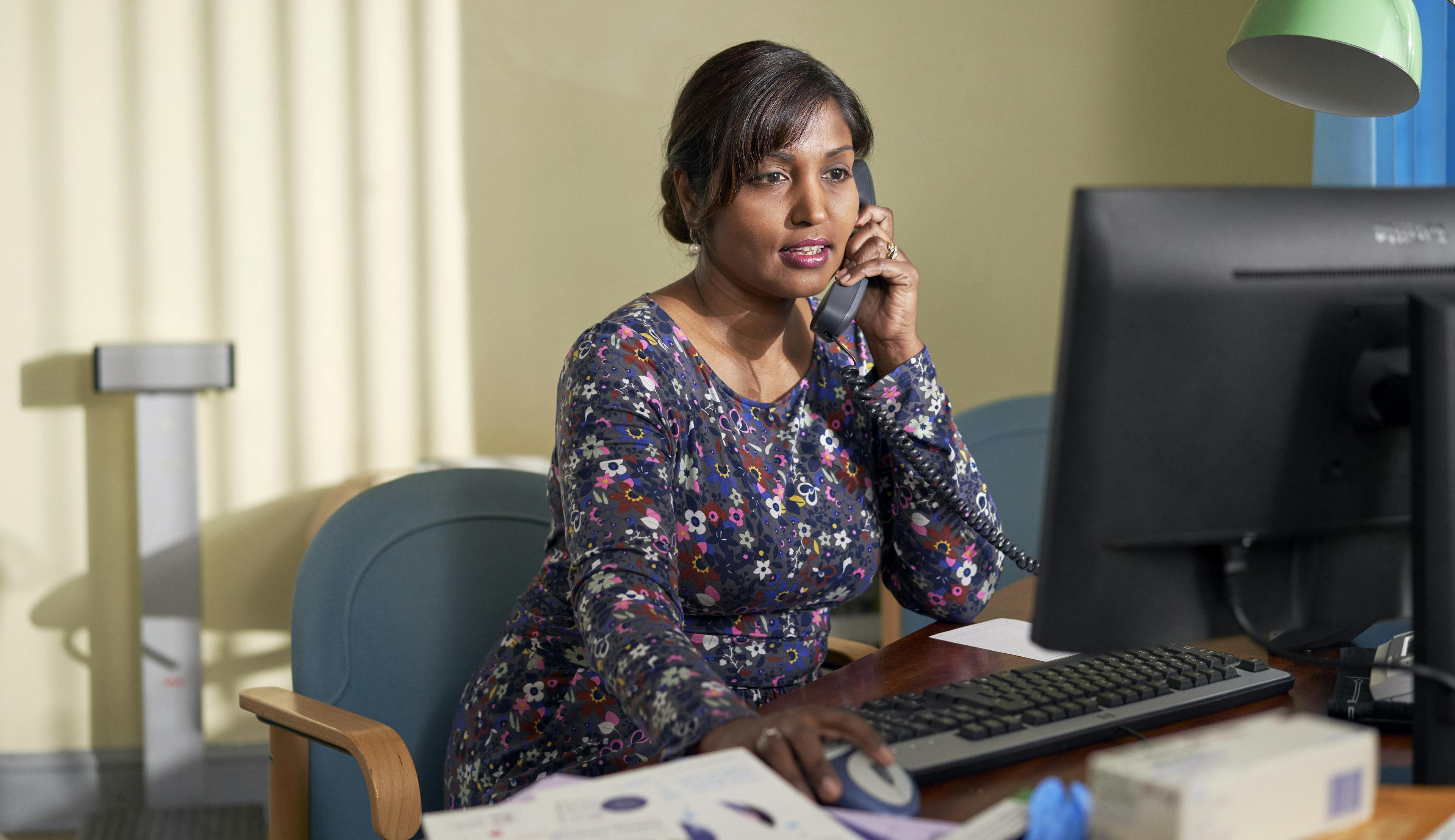 A GP is in her office, speaking on the phone and looking at her computer.